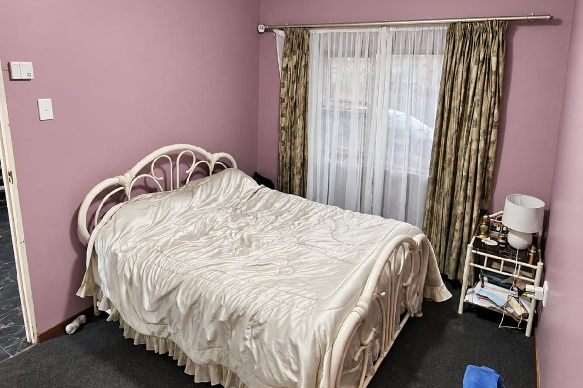 A bedroom featuring dark carpeting, mauve-toned walls, and a window dressed with curtains.