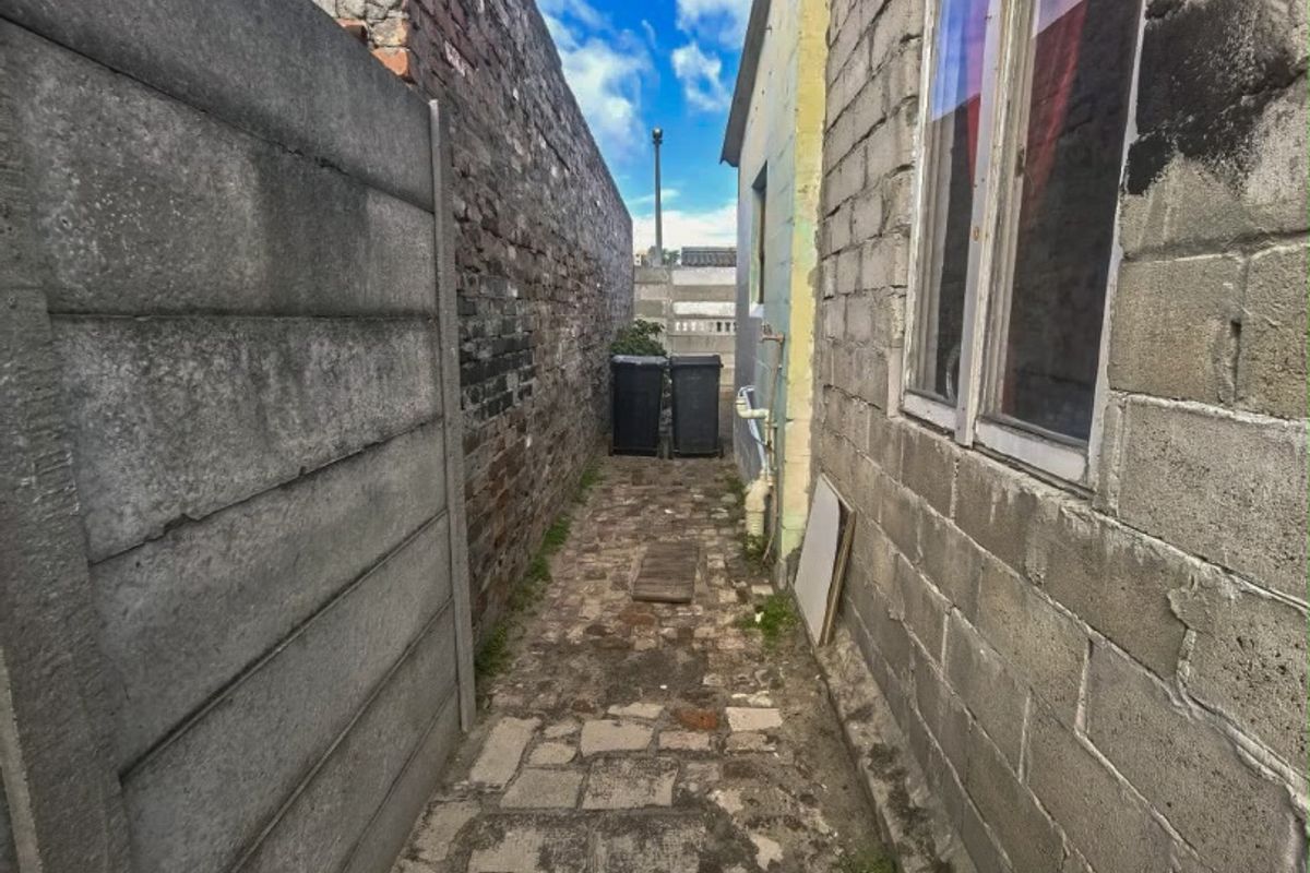 A narrow side passage featuring a stone paved walkway between an exposed brick wall and a precast concrete boundary wall.