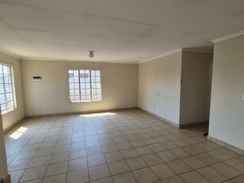 A spacious, empty living area featuring neutral tiled flooring, large windows with security bars, and a simple ceiling finish.