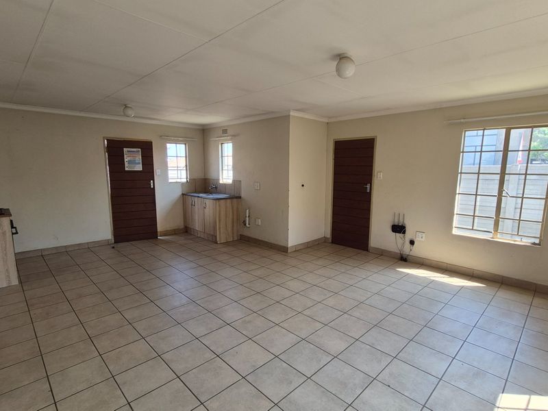 An open-plan living area featuring tiled flooring, a small kitchenette with built-in cabinetry, and natural light through multiple windows.