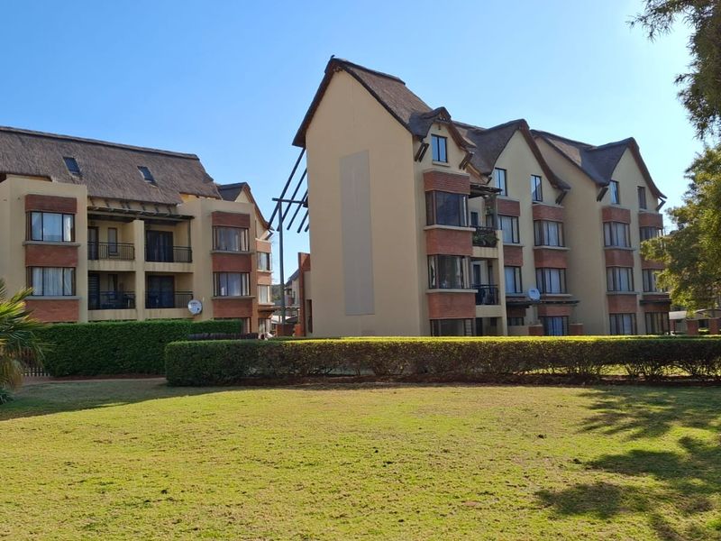 Exterior view of multi-story apartment buildings featuring thatched roofs, brick accents, and balconies overlooking a well-maintained communal lawn and garden area.