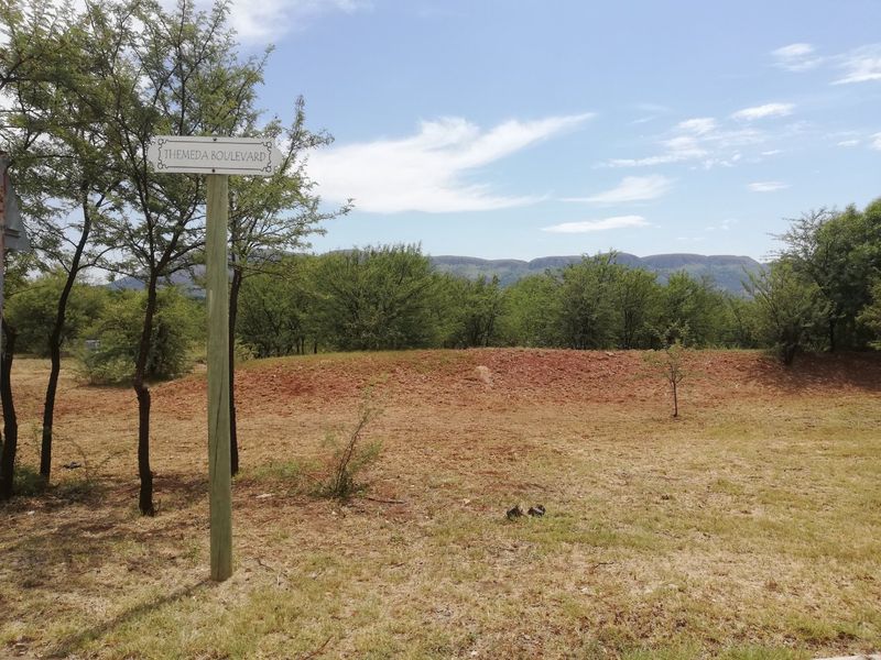 A view of the vacant residential land featuring native vegetation and a scenic mountain backdrop, marked by a street sign on Themeda Boulevard.