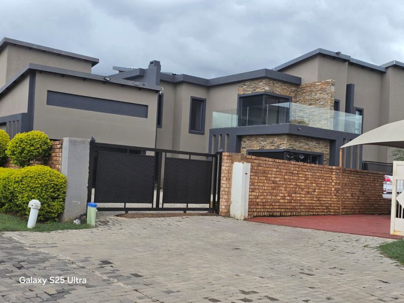 Modern double-storey residential property featuring stone cladding, a glass balcony, and a secure gated entrance with a paved driveway.