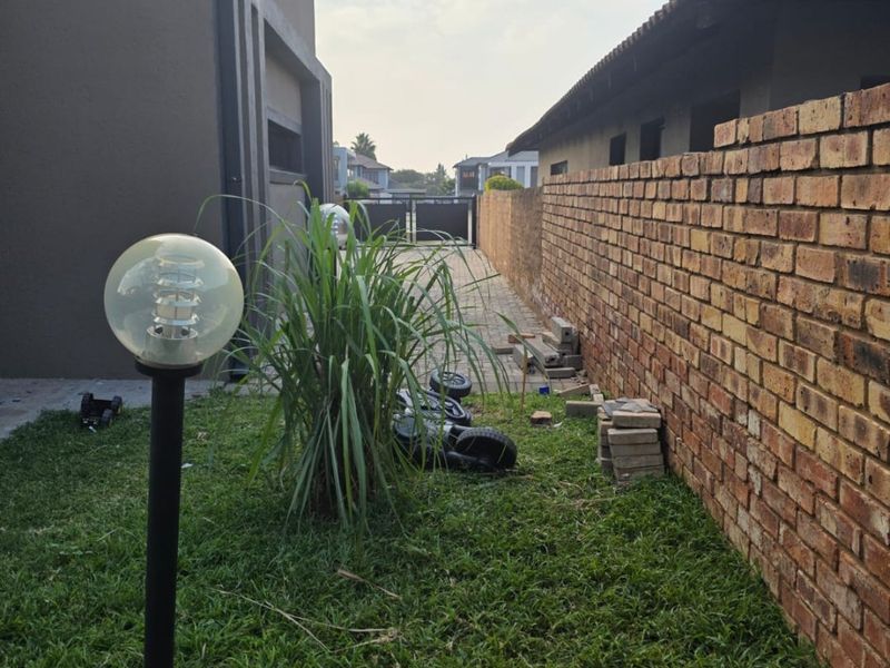 A side yard area featuring a brick boundary wall, paved driveway access, and a garden post light, currently showing signs of unfinished landscaping.
