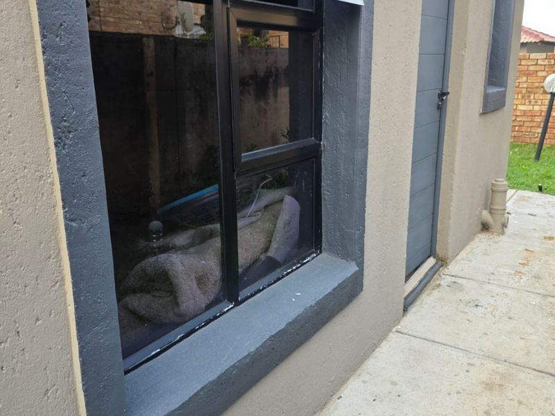 A view of the rear exterior wall featuring a dark-framed top-hung window, a side door, and a concrete walkway.
