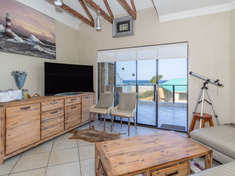 A bright living room featuring exposed timber ceiling beams, tiled flooring, and large sliding glass doors that open onto a balcony with ocean views.