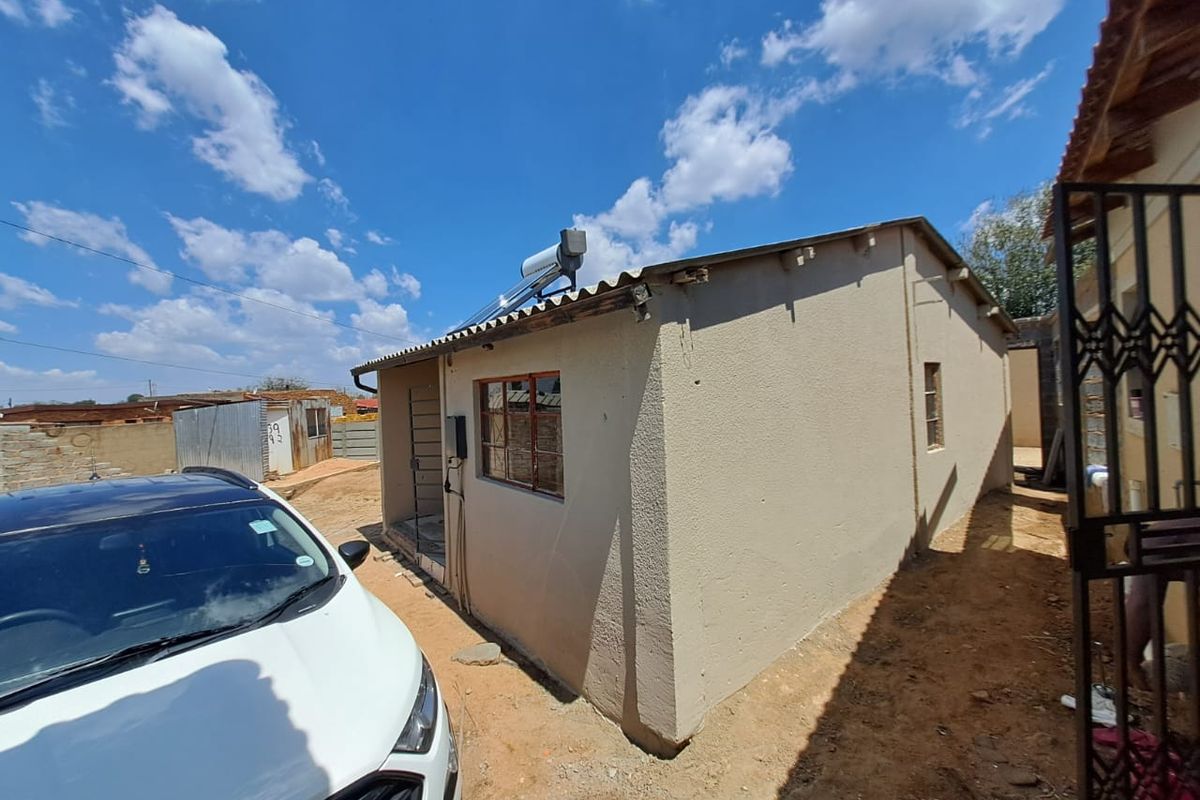 Rear exterior view of a single-story house featuring a solar water heater, corrugated roofing, and steel window frames with an unpaved yard area.