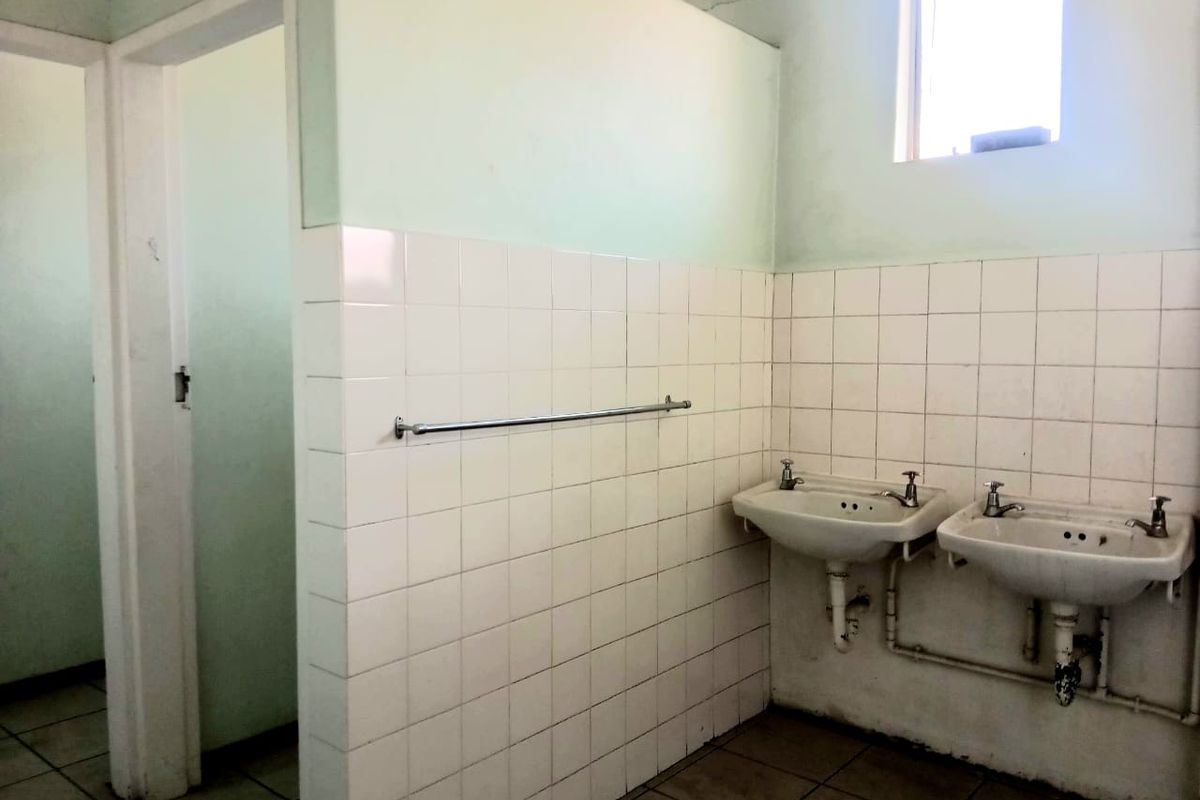 A functional commercial bathroom featuring dual wall-mounted basins, tiled splashbacks, and ceramic floor tiles, currently in need of cosmetic refurbishment.