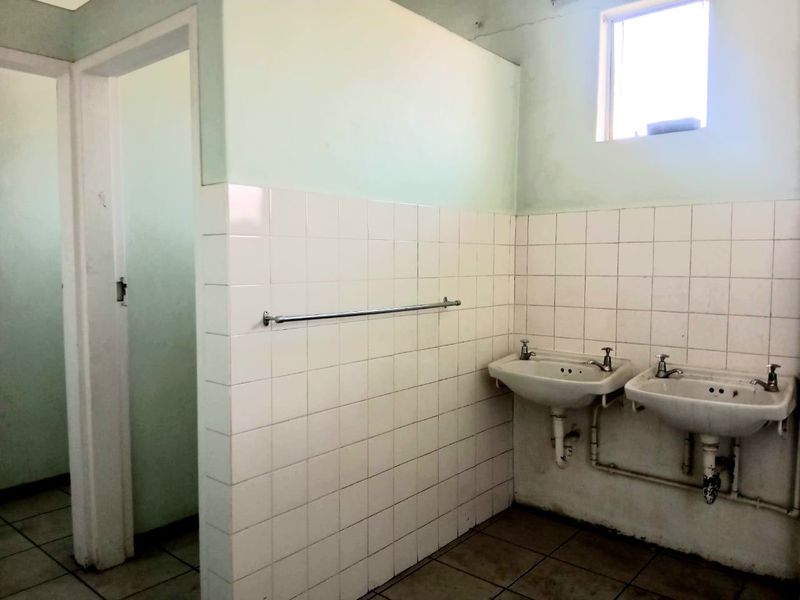 A functional commercial bathroom featuring dual wall-mounted basins, tiled splashbacks, and ceramic floor tiles, currently in need of cosmetic refurbishment.