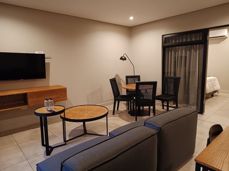 A modern open-plan living and dining area featuring neutral tiled flooring, recessed lighting, and a sliding glass door leading to an adjacent bedroom.