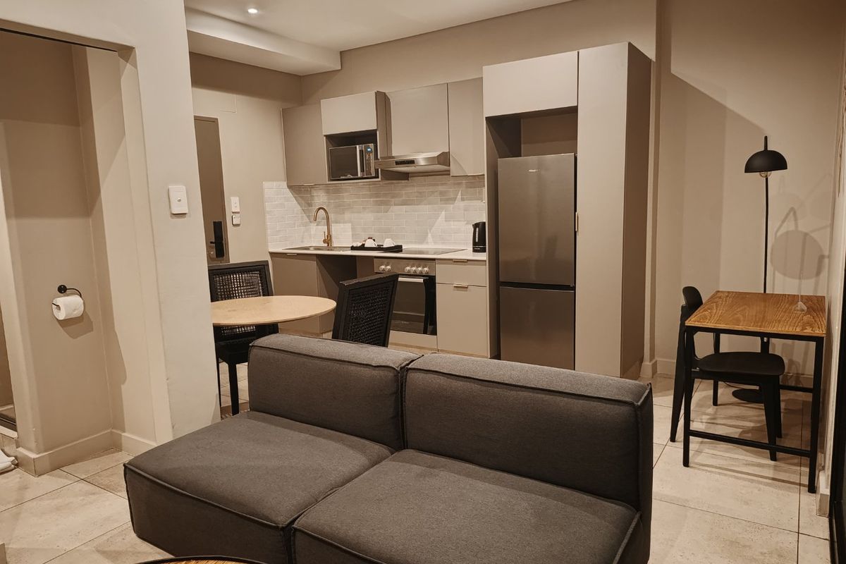 Modern open-plan apartment living area featuring a compact, well-equipped kitchen with built-in cabinetry, stainless steel appliances, and neutral tiled flooring throughout.