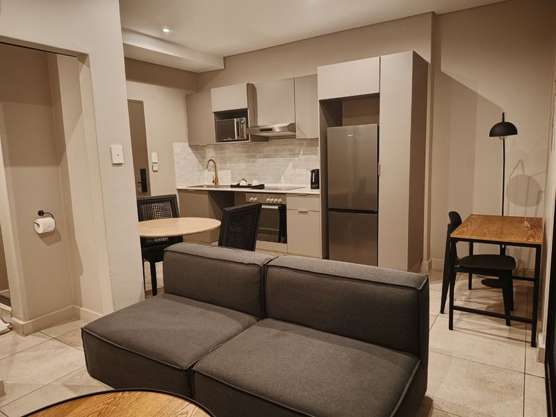 Modern open-plan apartment living area featuring a compact, well-equipped kitchen with built-in cabinetry, stainless steel appliances, and neutral tiled flooring throughout.