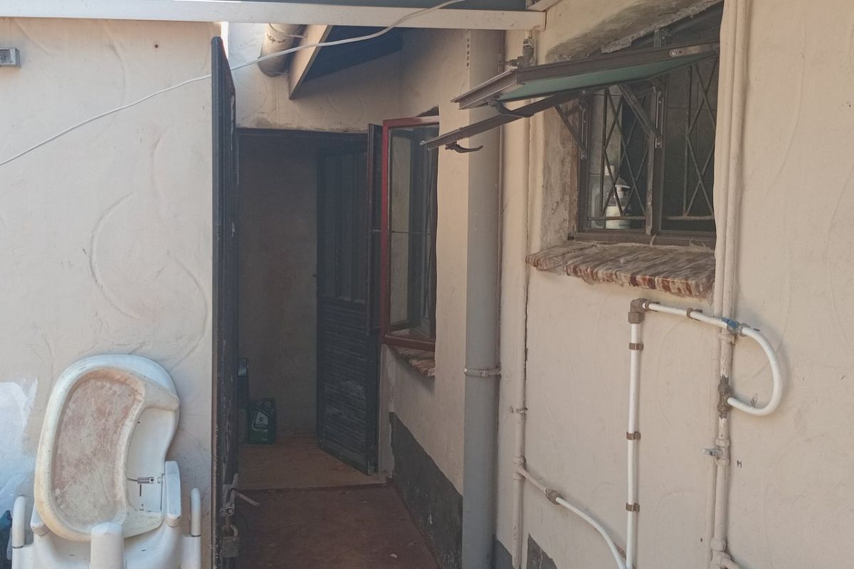 A narrow exterior rear passage area featuring textured walls, security gates, and exposed plumbing, showing signs of significant wear and weathering.