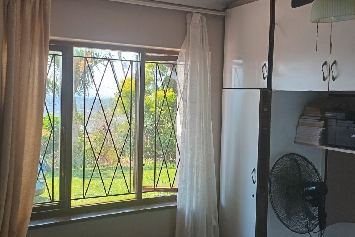 A bedroom featuring built-in cupboards and a window with security bars, overlooking the garden area.