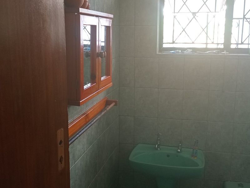 A dated bathroom featuring a pedestal basin, wall-mounted wooden cabinet, and tiled walls with security bars on the window.
