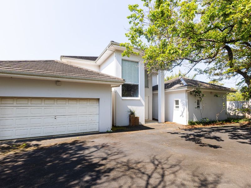 Front elevation of a residential property featuring a double garage, asphalt driveway, and mature trees providing shade to the building facade.