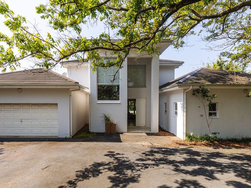 Front elevation of a modern house featuring a double garage, paved driveway, and a prominent central entrance under a mature tree canopy.