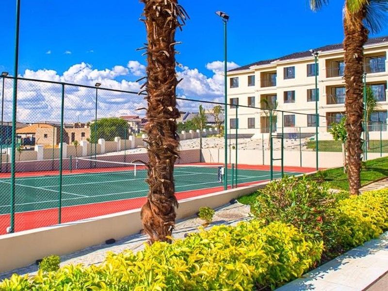 A well-maintained outdoor tennis court facility within the apartment complex, featuring perimeter fencing, floodlighting, and surrounding landscaped garden areas.