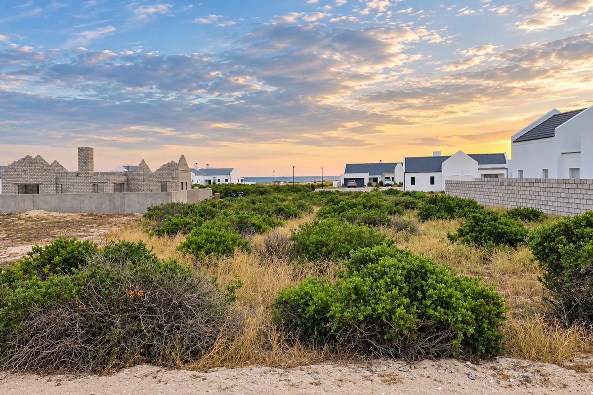 A vacant residential plot featuring natural coastal vegetation, surrounded by established residential properties and boundary walls in a quiet estate setting.