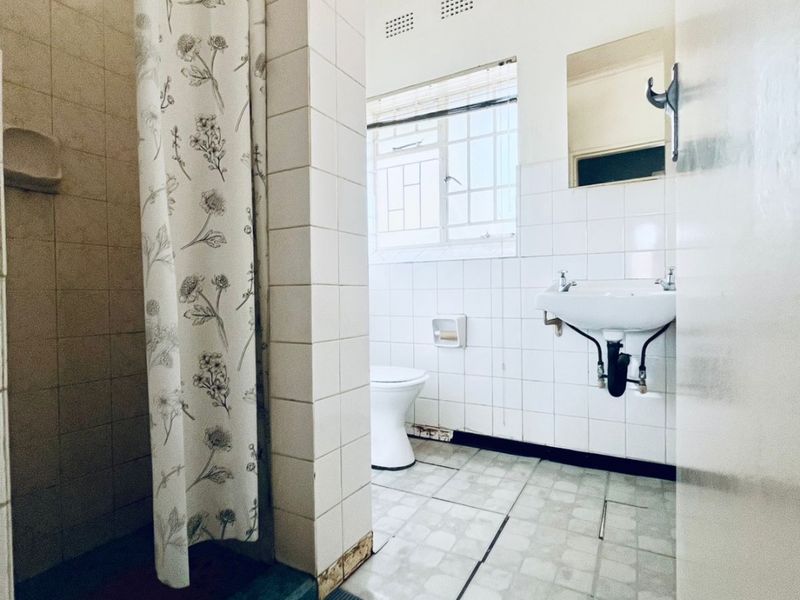 A functional bathroom featuring a shower, toilet, and wall-mounted basin with tiled finishes, requiring cosmetic updates to modernize the space.