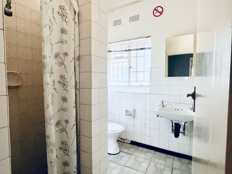 A functional bathroom featuring a shower, wall-mounted basin, and toilet, with tiled finishes throughout. The space is ready for modernization.