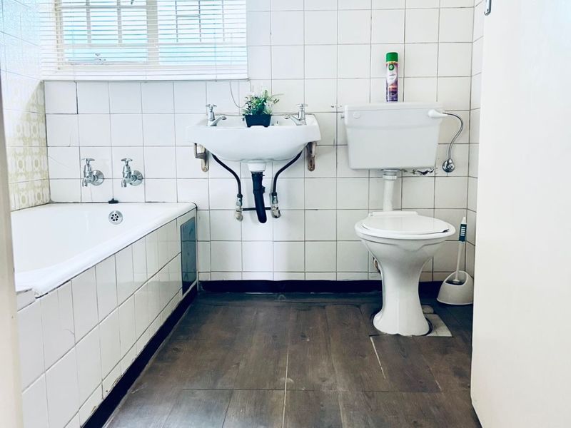 A dated bathroom featuring a bathtub, wall-mounted basin, and toilet with white ceramic wall tiles and wood-look laminate flooring.