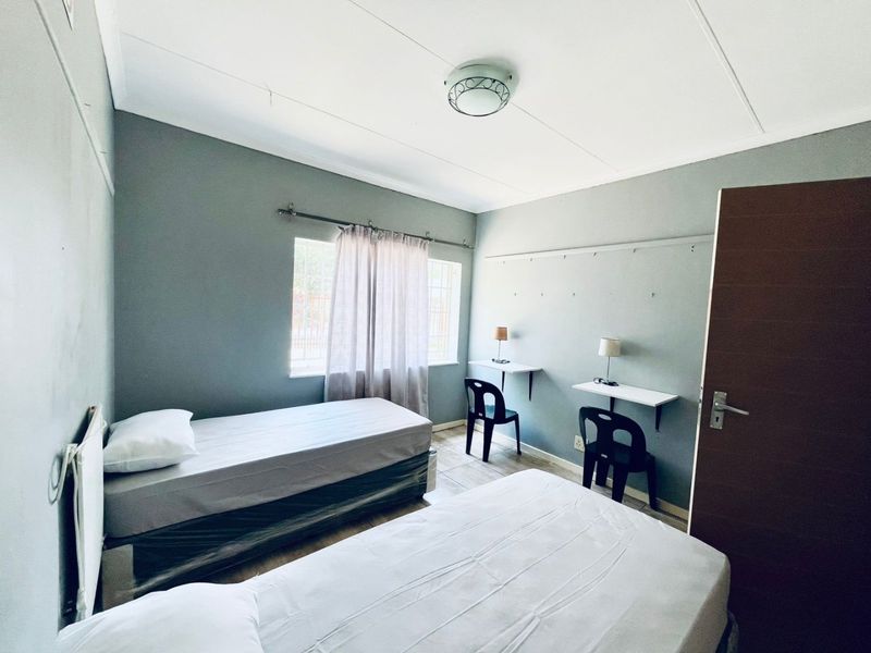 A functional bedroom featuring tiled flooring, built-in wall-mounted desks, and a window with security bars, offering a practical space for residents.