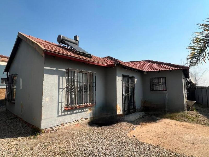 Rear view of a single-story house featuring a tiled roof, solar water heater, and security bars on windows, set on a gravel-covered yard.