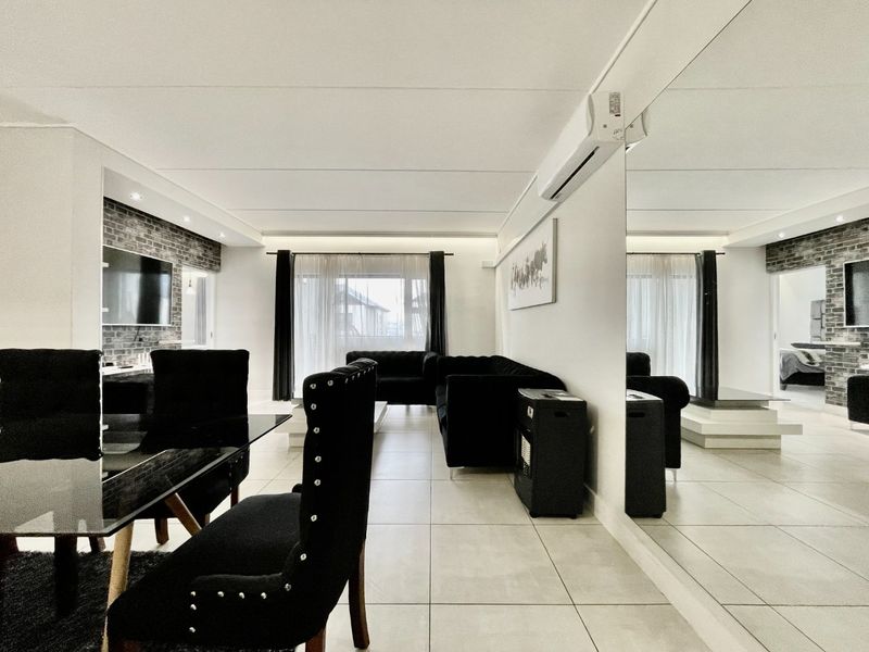 Spacious open-plan living and dining area featuring tiled floors, an exposed brick feature wall, recessed lighting, and a wall-mounted split-system air conditioner.