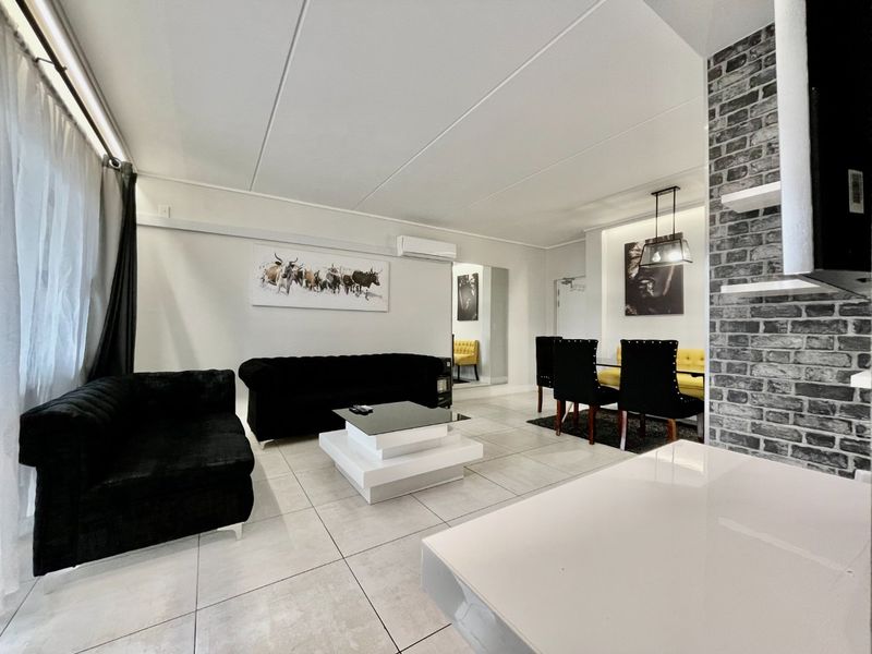 A bright open-plan living and dining area featuring modern tiled floors, a stylish exposed brick accent wall, and a wall-mounted air conditioning unit.