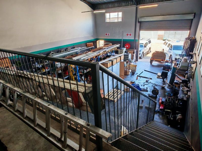 Industrial warehouse space featuring a mezzanine level with metal staircase, high ceilings with exposed steel trusses, and a large roller shutter door for access.