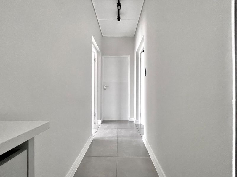 A clean, minimalist hallway featuring neutral tiled flooring, smooth walls, and modern track lighting, providing a bright and seamless transition between rooms.
