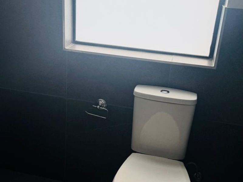 A clean, modern bathroom featuring dark floor-to-ceiling wall tiles, a white toilet, and a top-hung window providing natural light.