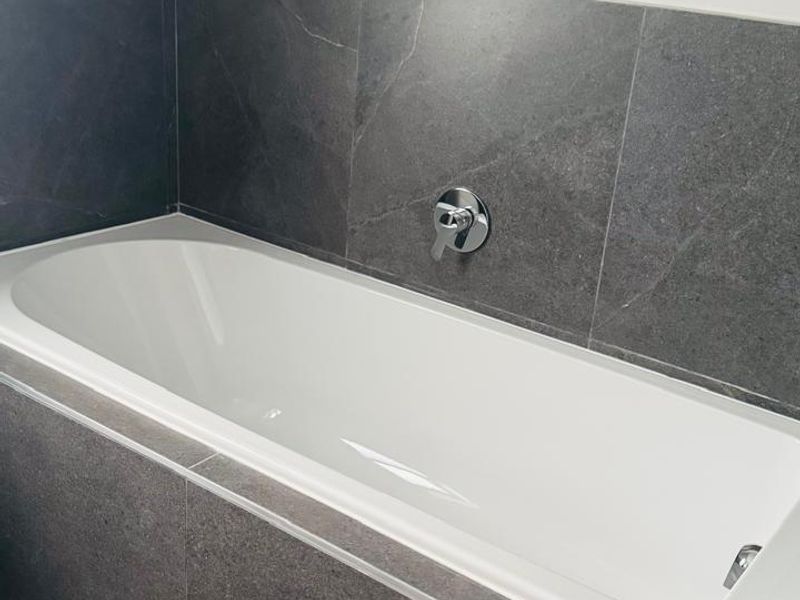 A modern bathroom featuring a clean built-in bathtub with dark grey stone-look wall tiling and a wall-mounted chrome mixer tap.