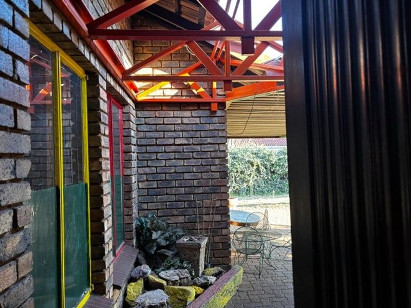 A covered outdoor patio area featuring exposed brick walls, steel roof trusses, and a built-in planter box, leading out to a paved garden space.