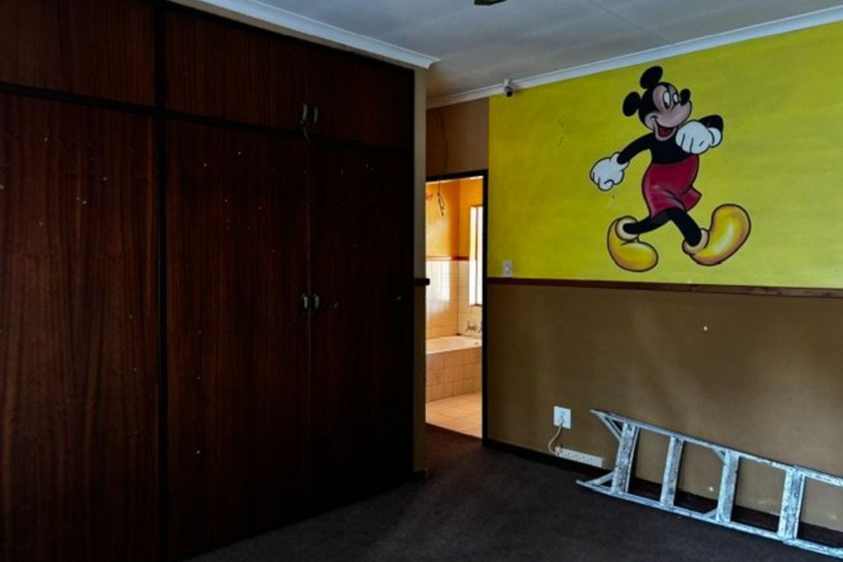 A bedroom featuring built-in wooden cupboards and dated wall finishes, with an adjacent view into a bathroom area.