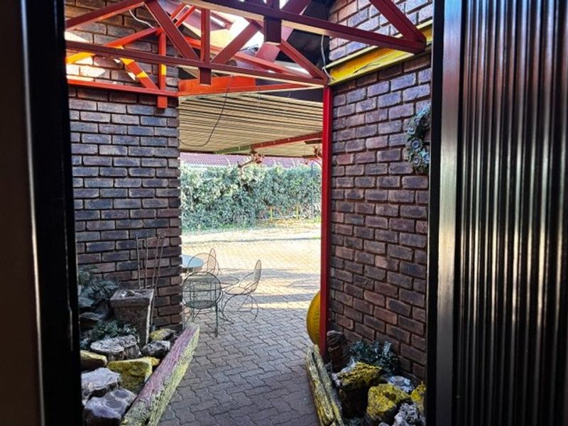 Covered outdoor patio area featuring exposed brick walls and industrial-style steel roof trusses, leading out to a paved garden space.