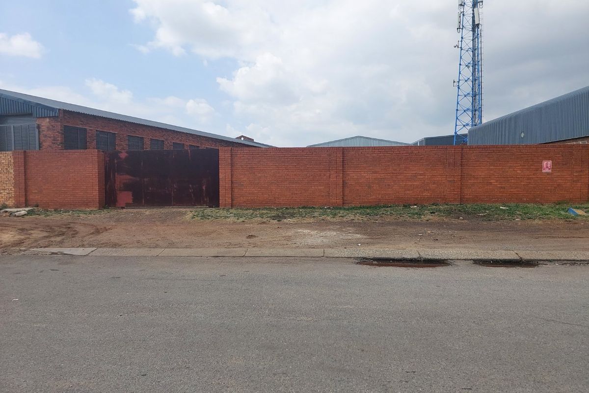 Street-level view of a commercial property featuring a brick perimeter wall, metal gate, and industrial structures with an adjacent telecommunications tower.