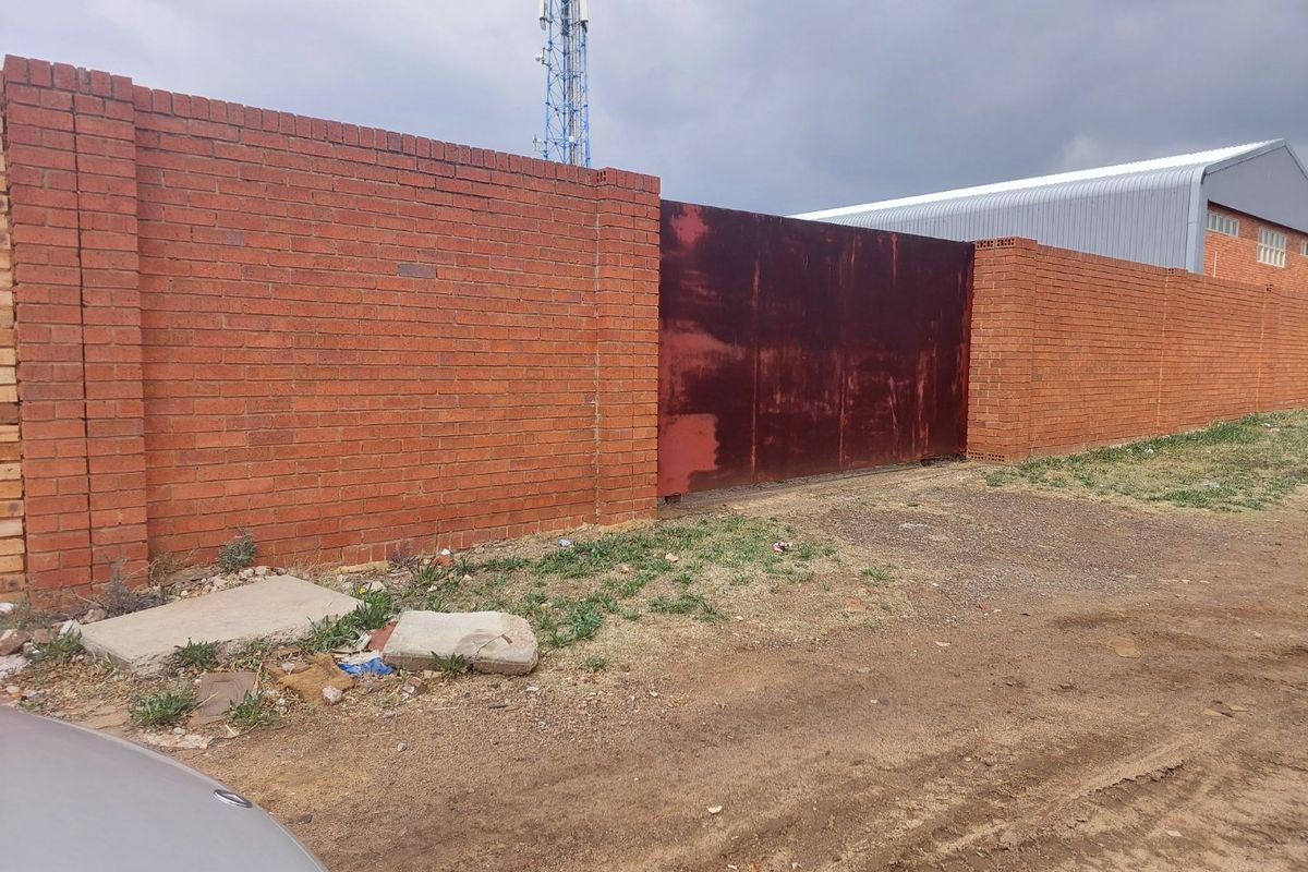 Front view of a vacant residential plot featuring a brick perimeter wall and a large, weathered metal sliding gate.