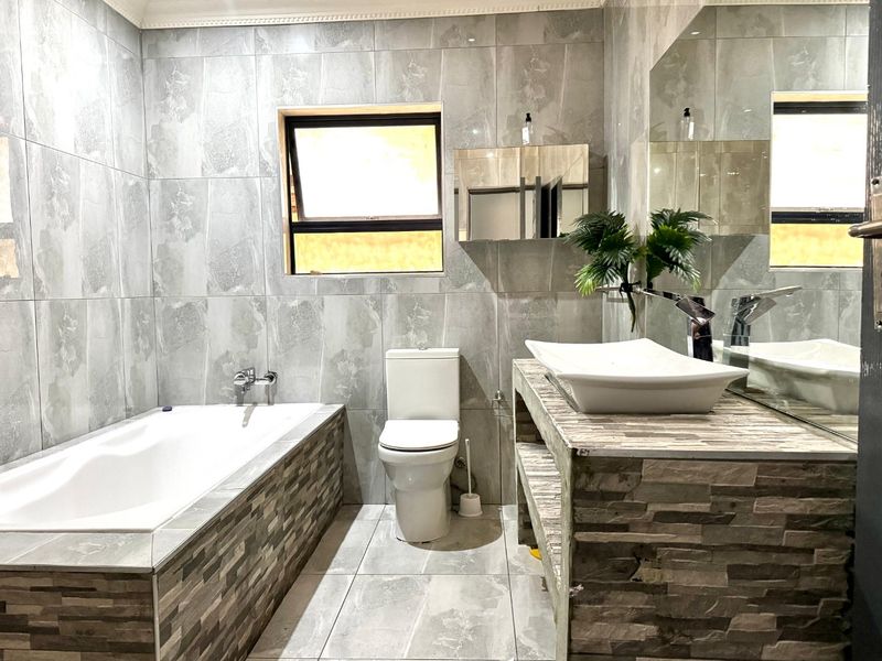 Modern bathroom featuring full-height grey tiling, a built-in bathtub, a stone-clad vanity with a vessel sink, and a wall-mounted mirror.