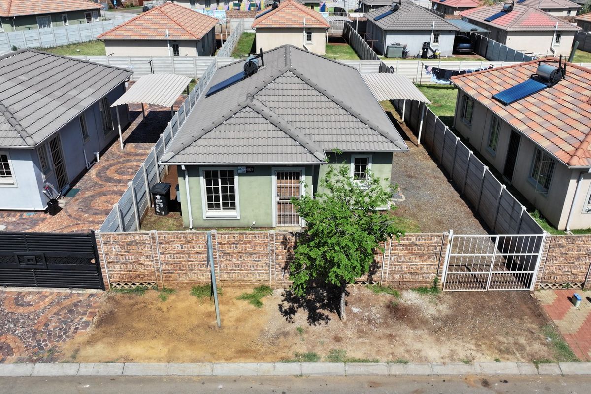 Aerial view of a detached residential house featuring a tiled roof, solar water heater, paved driveway, carport, and enclosed perimeter walling.