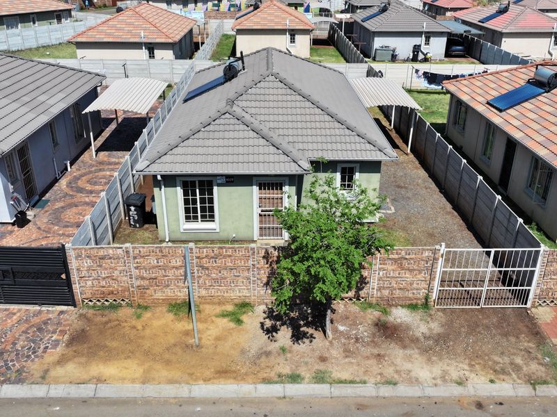 Aerial view of a detached residential house featuring a tiled roof, solar water heater, paved driveway, carport, and enclosed perimeter walling.