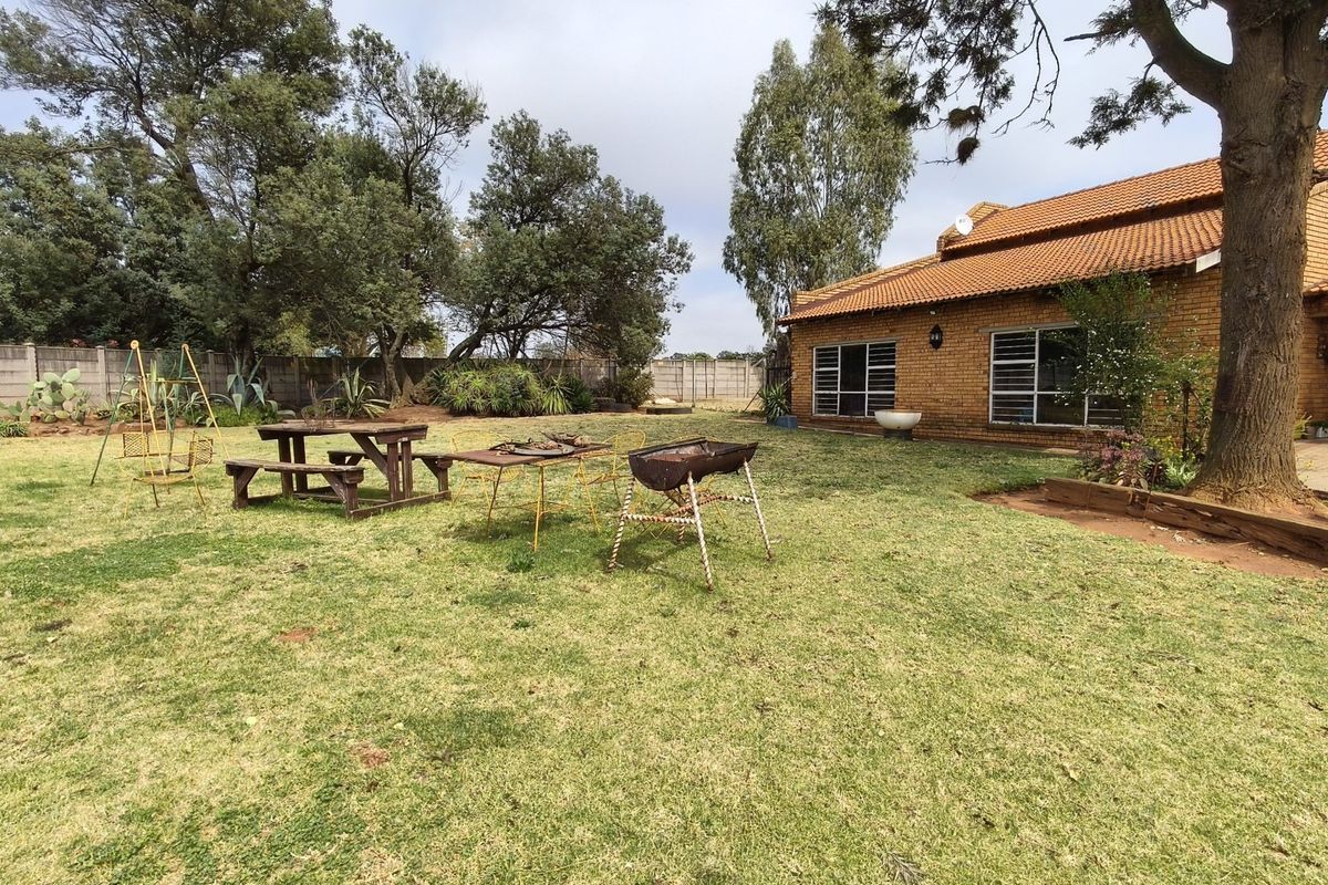 A spacious rear garden featuring a brick-built farmhouse with a tiled roof, mature trees, and a concrete boundary wall.