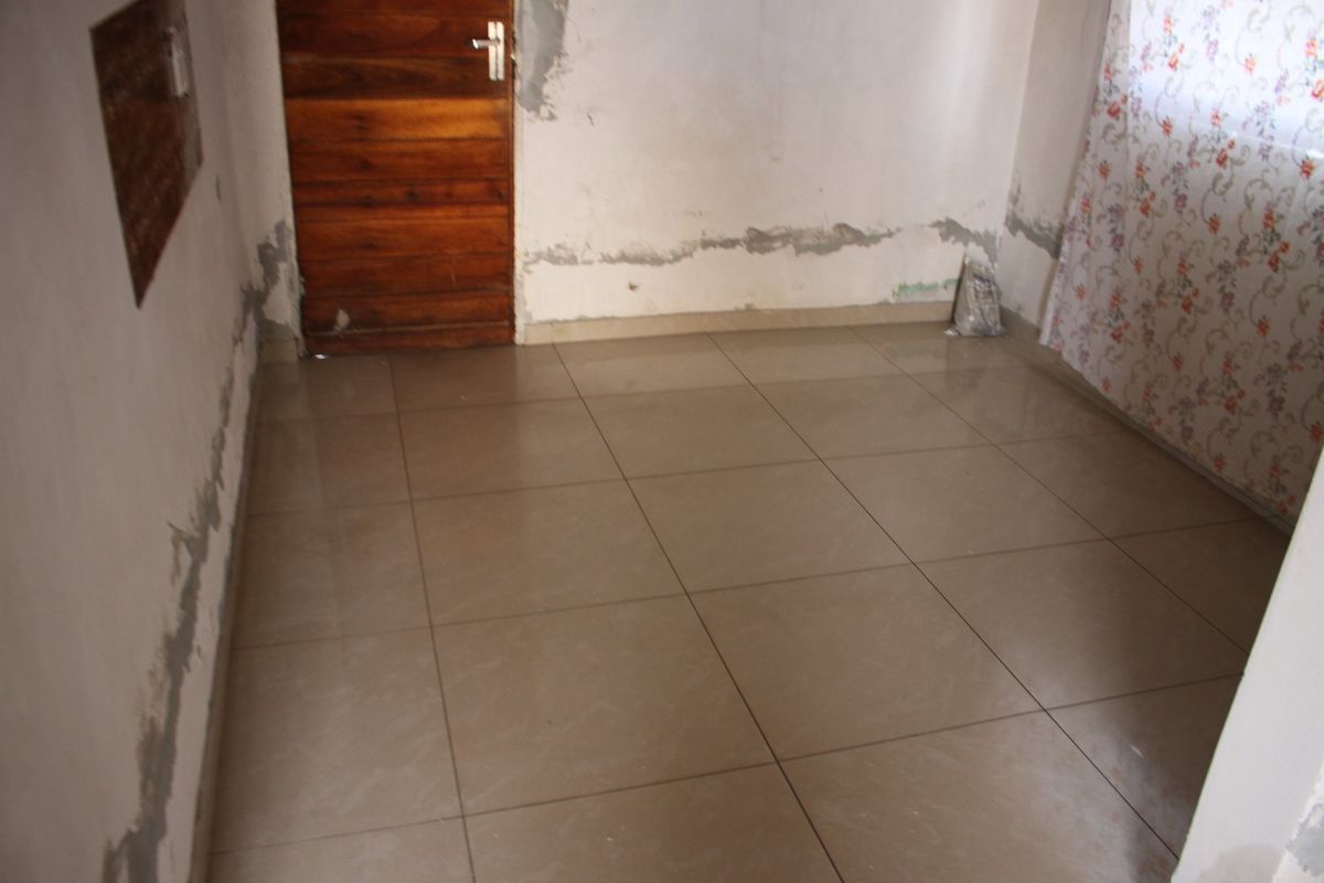 An empty bedroom featuring tiled flooring and a wooden door, currently requiring wall repairs and finishing to improve its overall condition.