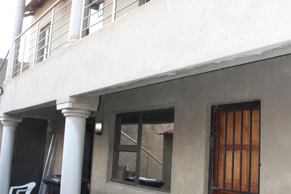 A two-story exterior view showing a ground floor patio with tiled flooring, concrete columns, and an upper-level balcony with a metal balustrade.