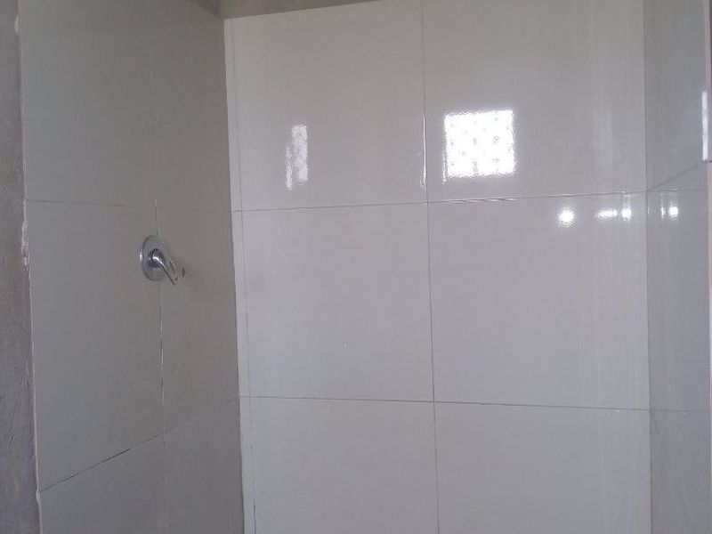 A shower enclosure featuring white wall tiles, mosaic floor tiling, and an electric shower head, with unfinished plaster walls above the tiled area.