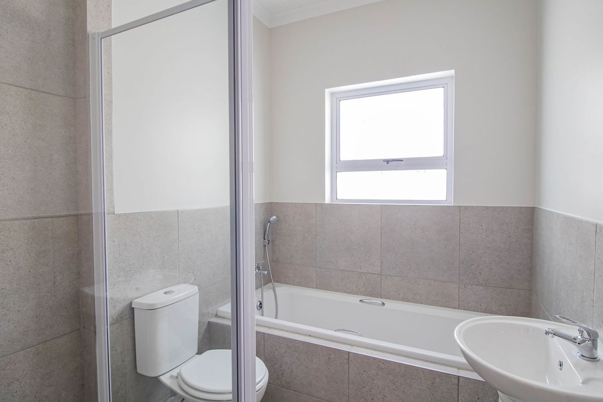A clean, modern bathroom featuring a built-in bathtub, glass shower screen, pedestal basin, and neutral wall tiling.