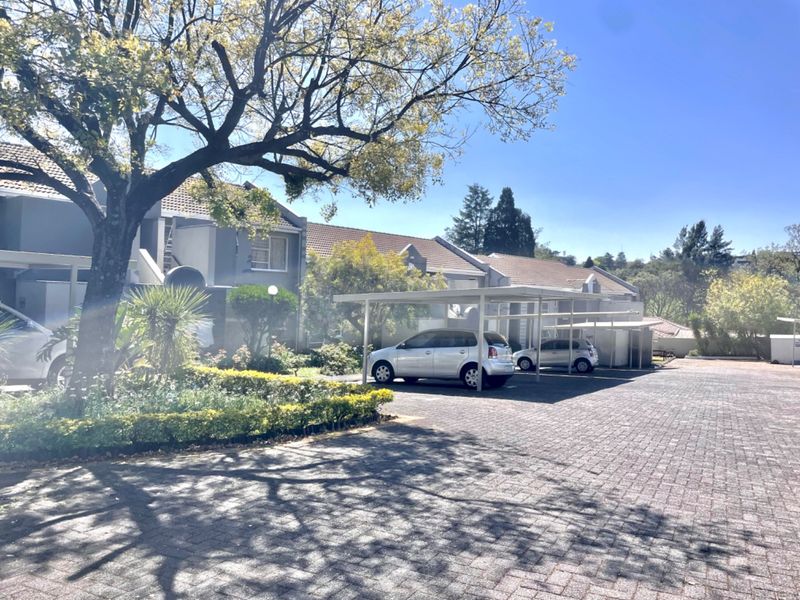 Front exterior view of the townhouse complex featuring a paved driveway, covered carports, and established garden landscaping under a clear sky.