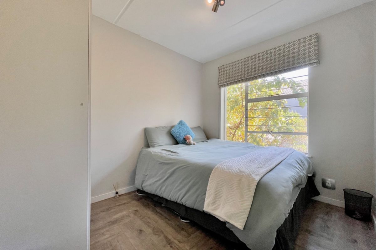 A compact bedroom featuring laminate flooring, a built-in cupboard, and a large window with a roller blind, offering natural light and garden views.