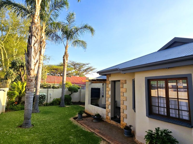 A well-maintained rear garden featuring a paved walkway, built-in braai area, timber pergola, and established palm trees against a plastered exterior wall.